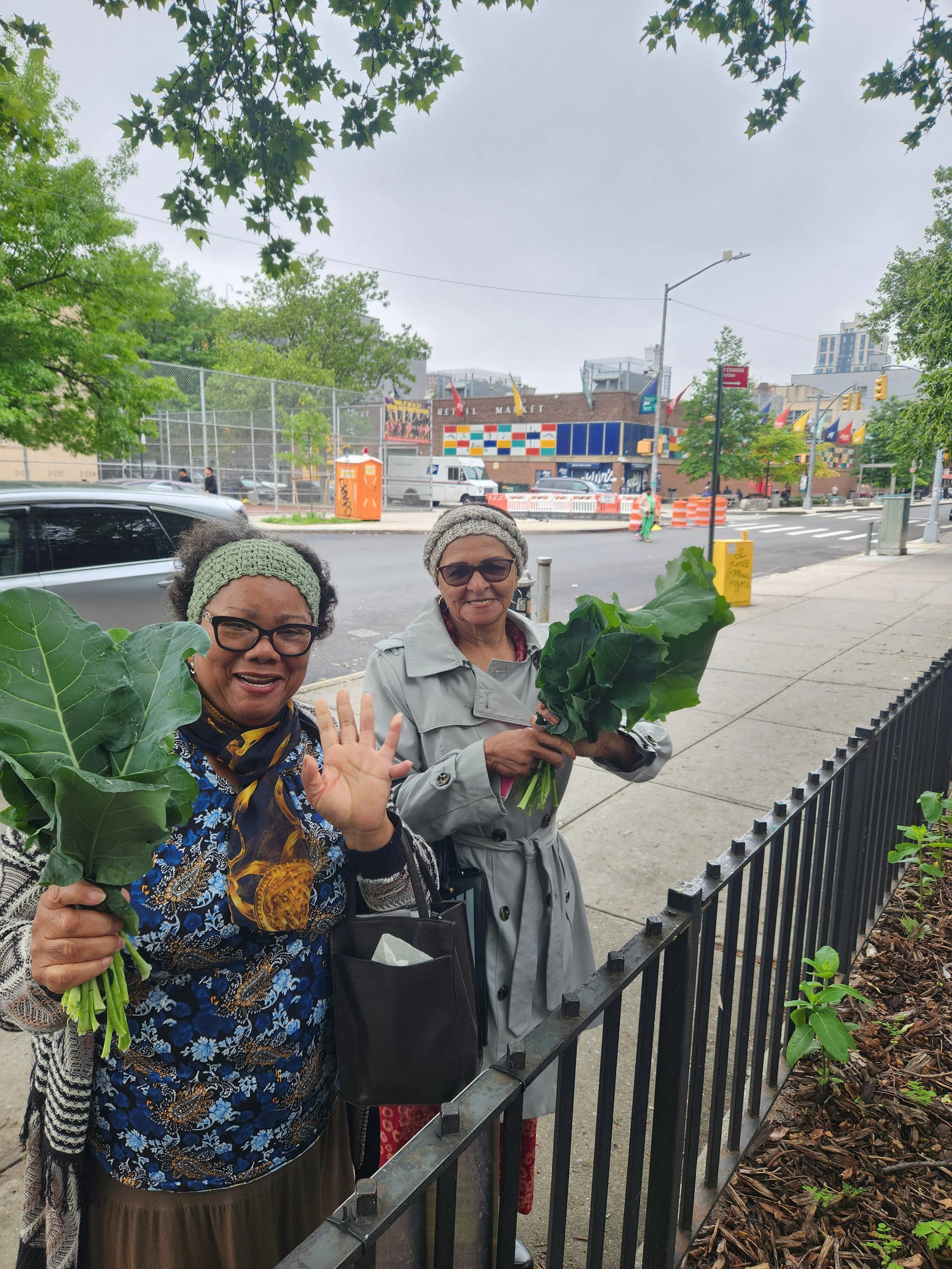 Two women standing on a city sidewalk holding leafy greens, smiling and waving at the camera.
