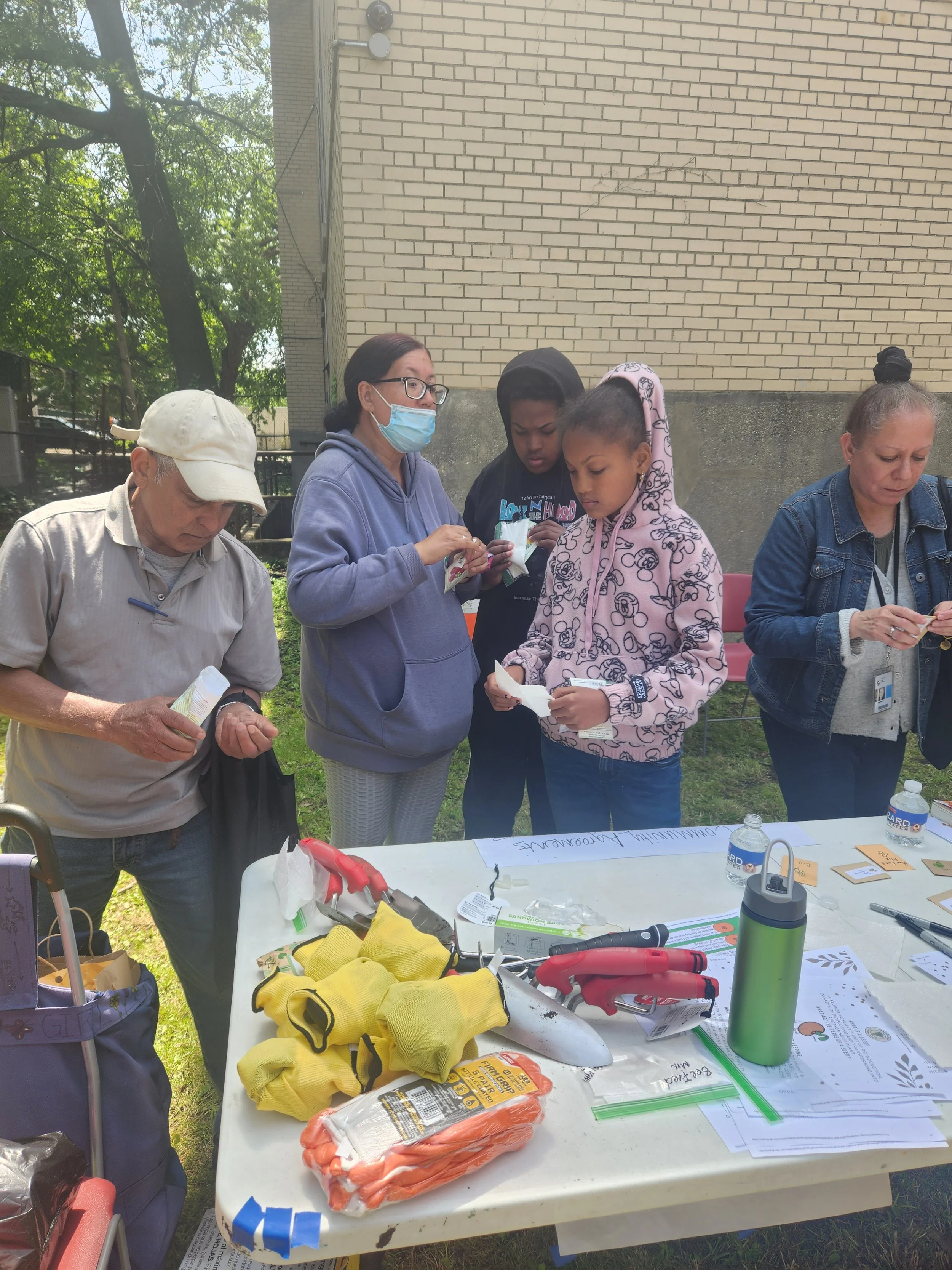 Group of five people standing outside at a table, examining items, with gardening tools, water bottles, and papers on the table.