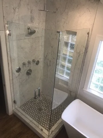 Empty glass shower with marble walls and a small tiled floor, adjacent to a window and a white bathtub.