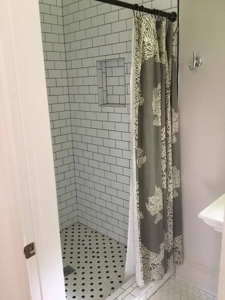 A walk-in shower with white subway tile walls, a built-in niche, a black and white hexagon tiled floor, and a patterned shower curtain.