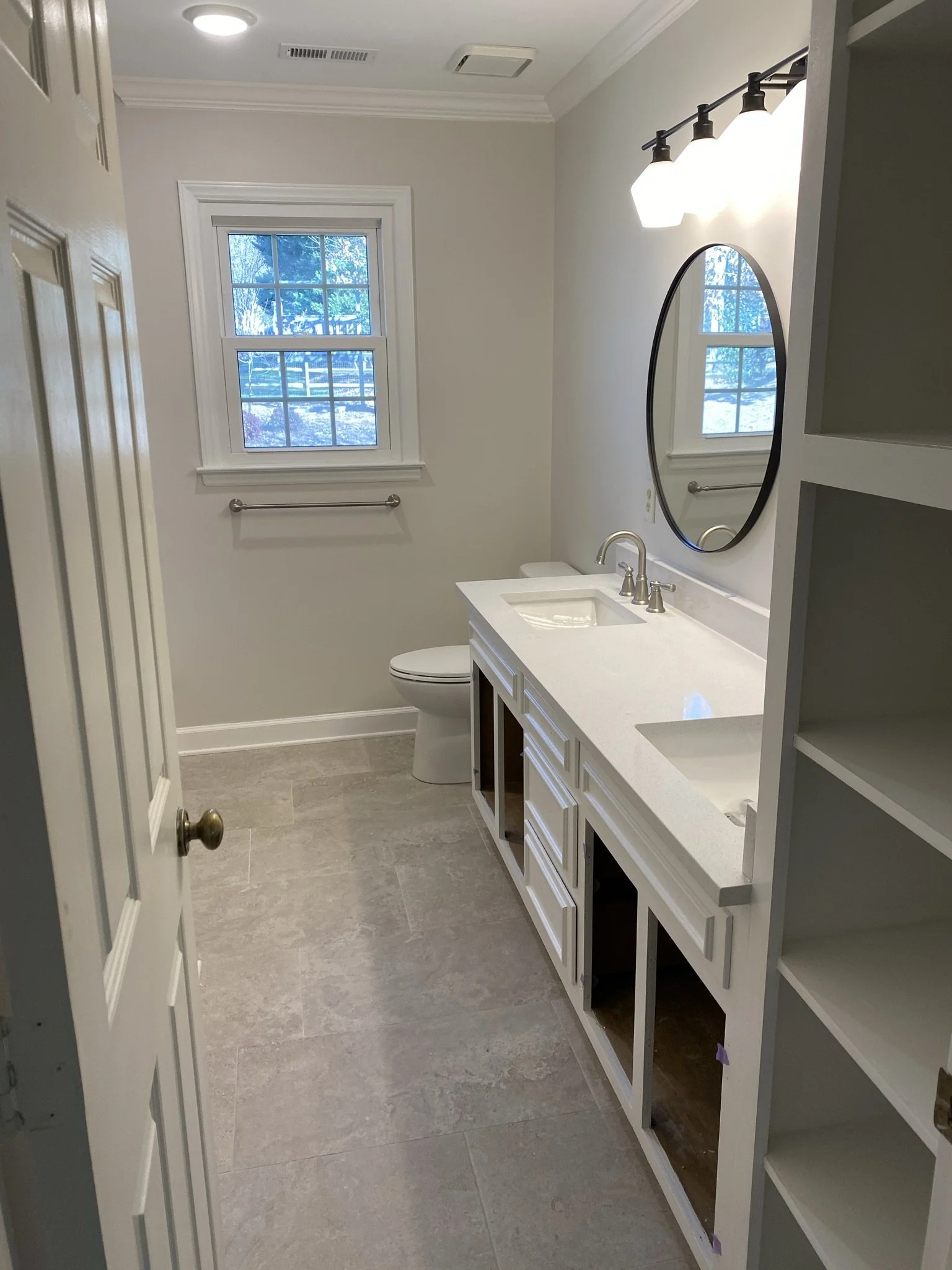 Bathroom with unfinished white cabinet and shelves, oval mirror, window with outdoor view of trees and snow, ceiling light fixture, tile flooring, and a white toilet.