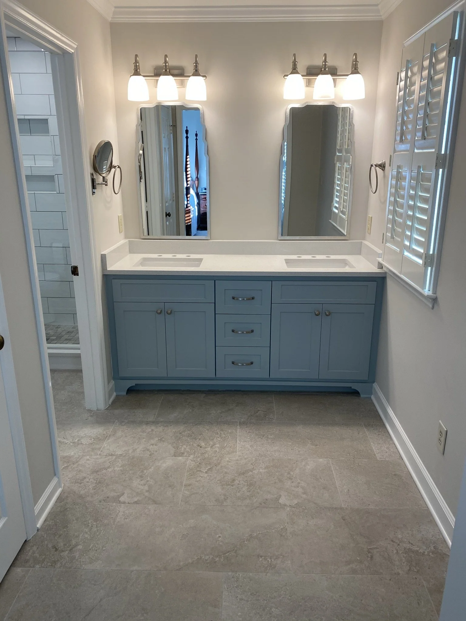 Bathroom vanity with a large mirror, two sinks, and overhead lighting fixtures; visible shower area with white tiles and glass door; neutral-colored walls and tile flooring.