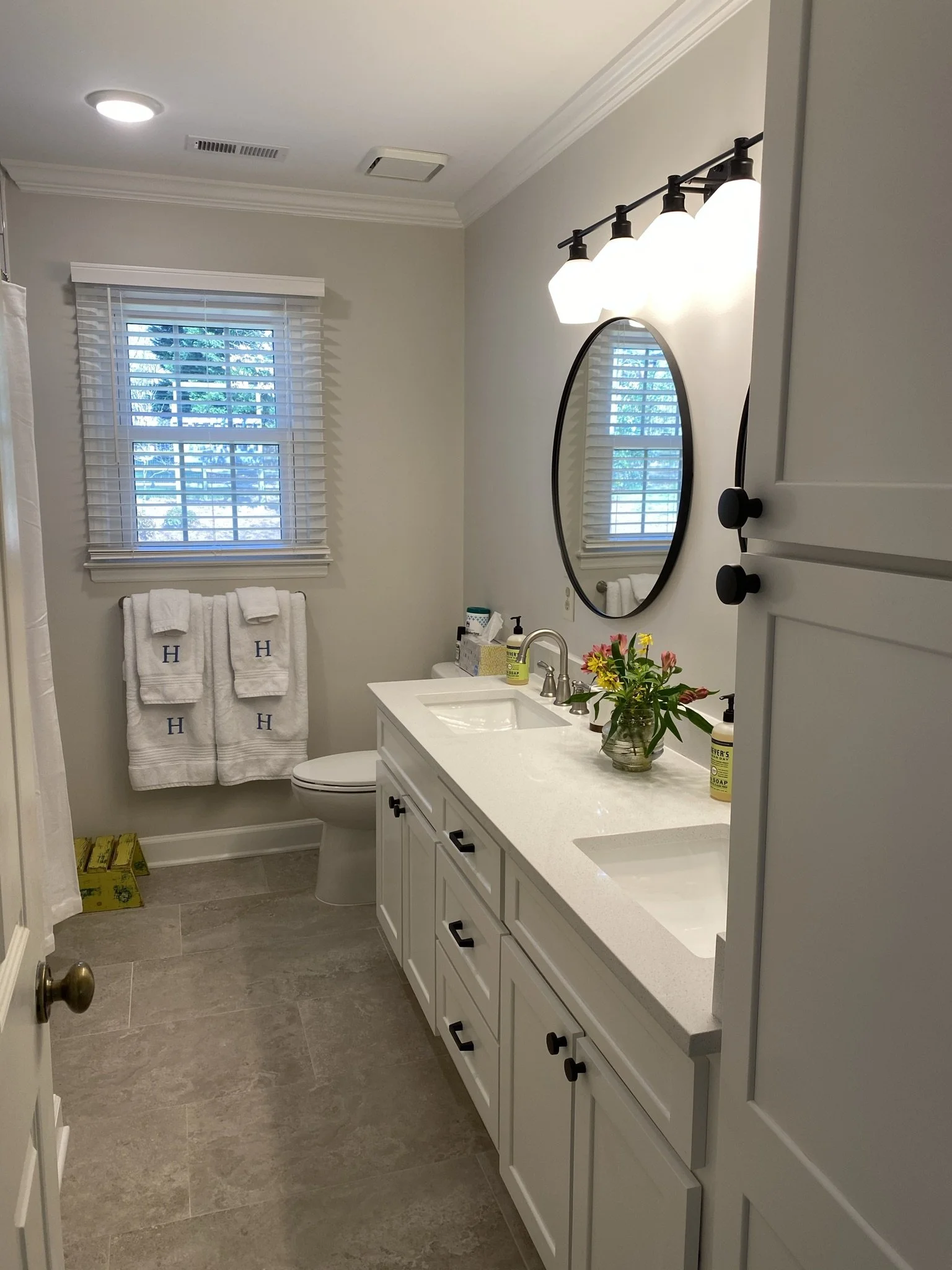 A bathroom with a white vanity, a round mirror, a tiled floor, a window with white blinds, a towel rack with towels monogrammed with H, a vase of flowers, and bathroom essentials on the countertop.
