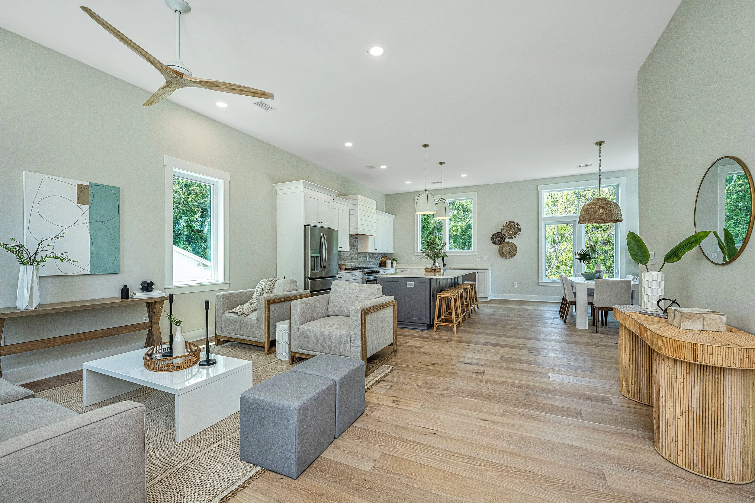 Open-concept living room and kitchen with modern furniture, hardwood floors, and large windows with greenery outside.