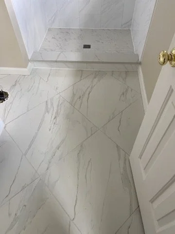 Bathroom floor with white marble tiles and a shower area with similar tiles, a drain, and a closed door.