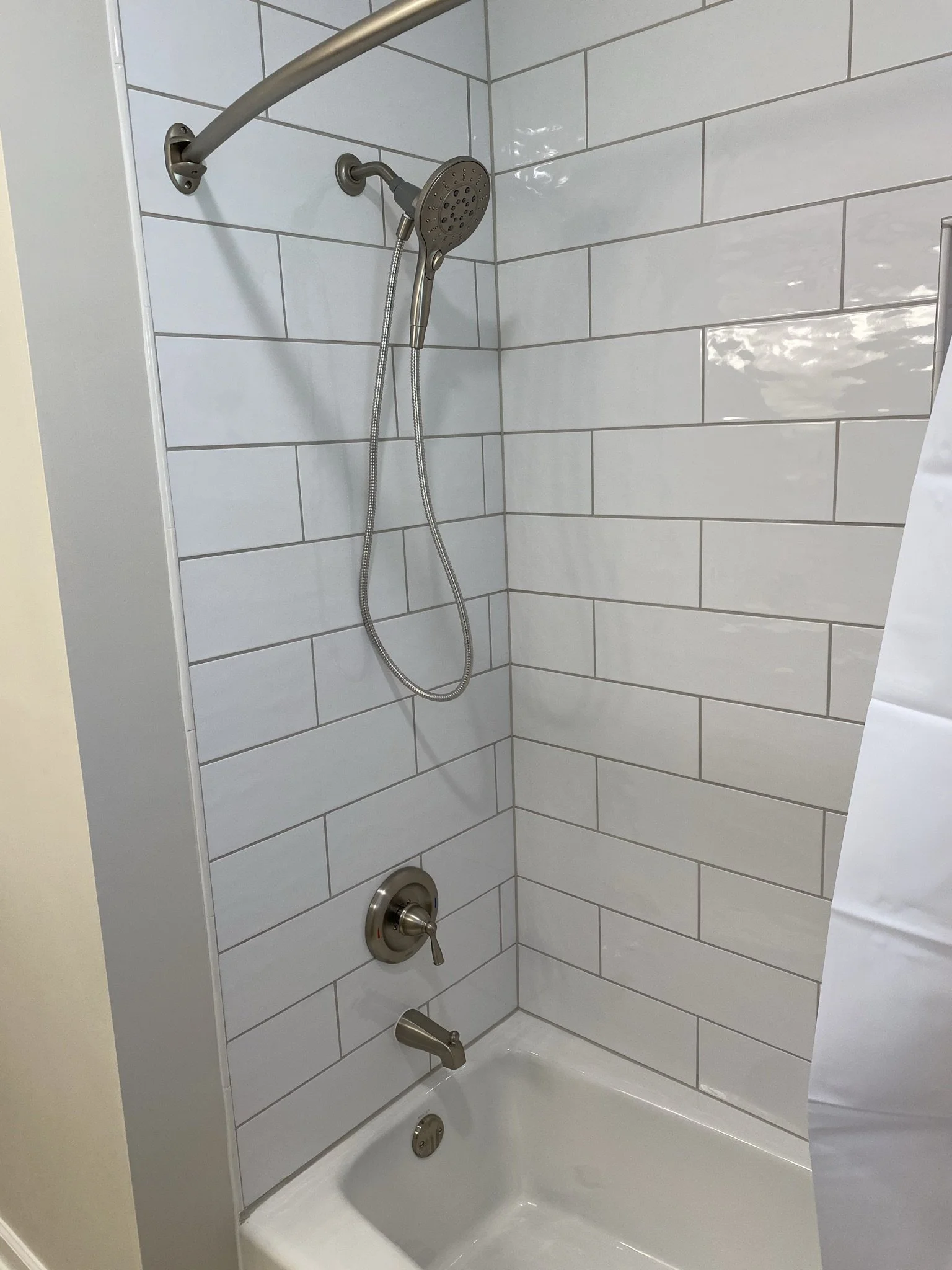 Shower with tiled walls, a handheld showerhead, a bathtub faucet, and a bathtub drain, all in stainless steel finish.
