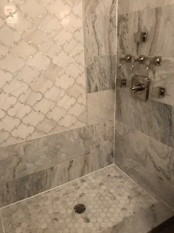 A tiled shower with gray and white marble tiles, a small drain on the floor, and multiple shower fixtures.