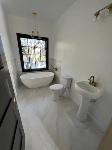 Bathroom with a freestanding bathtub, toilet, and pedestal sink, window with a black frame, marble floor, and simple lighting fixtures.