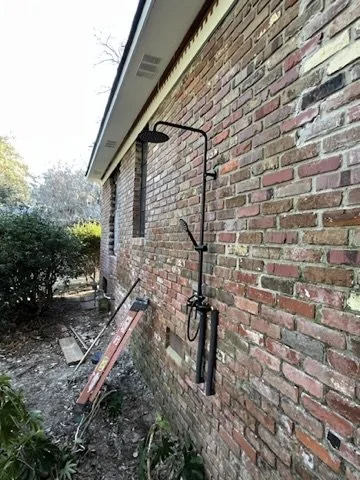 An outdoor shower mounted on a red brick house wall, with a garden and bushes nearby.