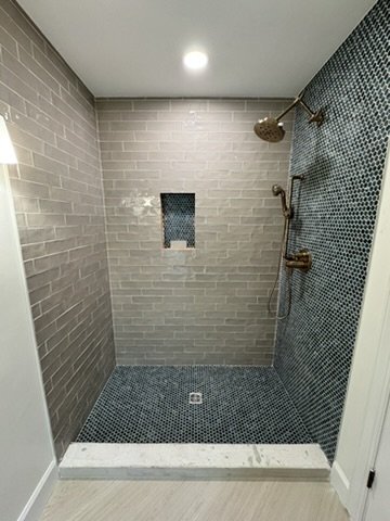 A walk-in shower with gray brick-like tiles on the walls, small blue mosaic tiles on the floor, and a built-in niche for toiletries. It features a rainfall showerhead, a hand-held shower, and a wall-mounted control.