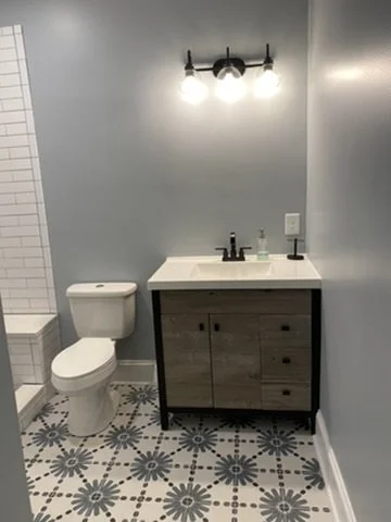 Bathroom with a toilet, wooden vanity with a sink, wall-mounted light fixture, gray wall, and patterned tile floor.
