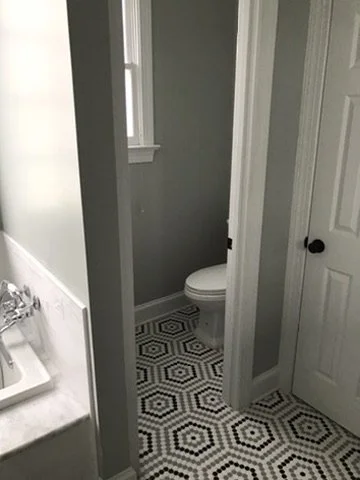 Small bathroom with a toilet, a window, a closed door, and black and white hexagonal patterned floor tiles.