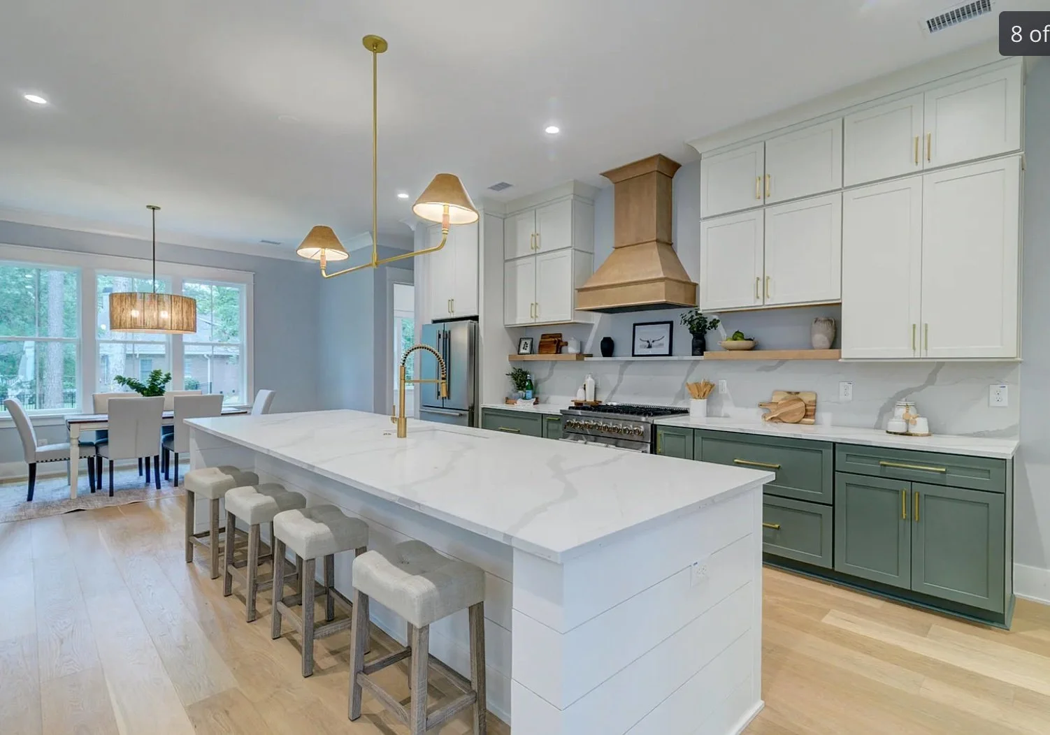 Modern kitchen with a large white island, gold faucet, green lower cabinets, white upper cabinets, gold hardware, a gold vent hood, and a dining area with large windows and pendant lighting.