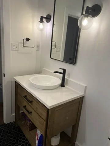 Bathroom sink with a wooden cabinet, white countertop, round vessel sink, black faucet, wall mirror, and wall-mounted light fixtures.