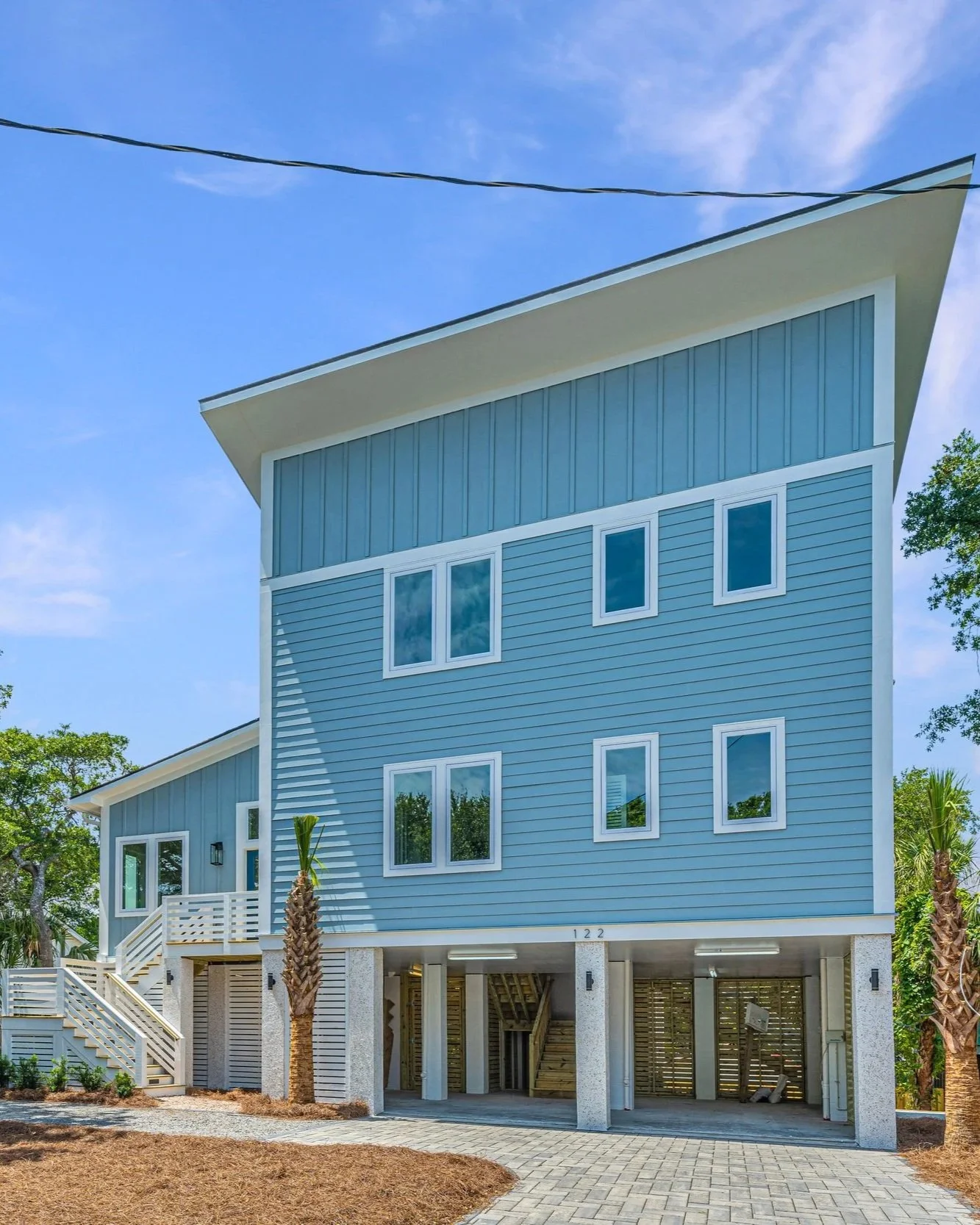 Blue modern multi-story house with large windows and a sloped roof, situated on a sandy lot with a paved driveway and palm trees. Under construction or renovation.
