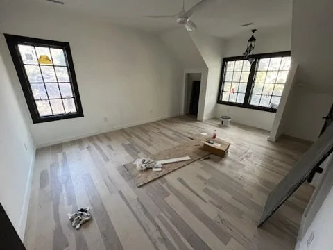 An unfinished room with light wood flooring, two large windows with black frames, a ceiling fan, and a hanging light fixture. Construction tools and materials are scattered on the floor.