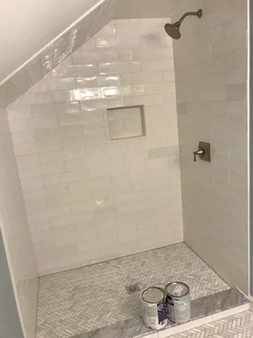 Empty shower with white subway tiles, a built-in niche, a ceiling shower head, and two cans of paint on the tiled floor.