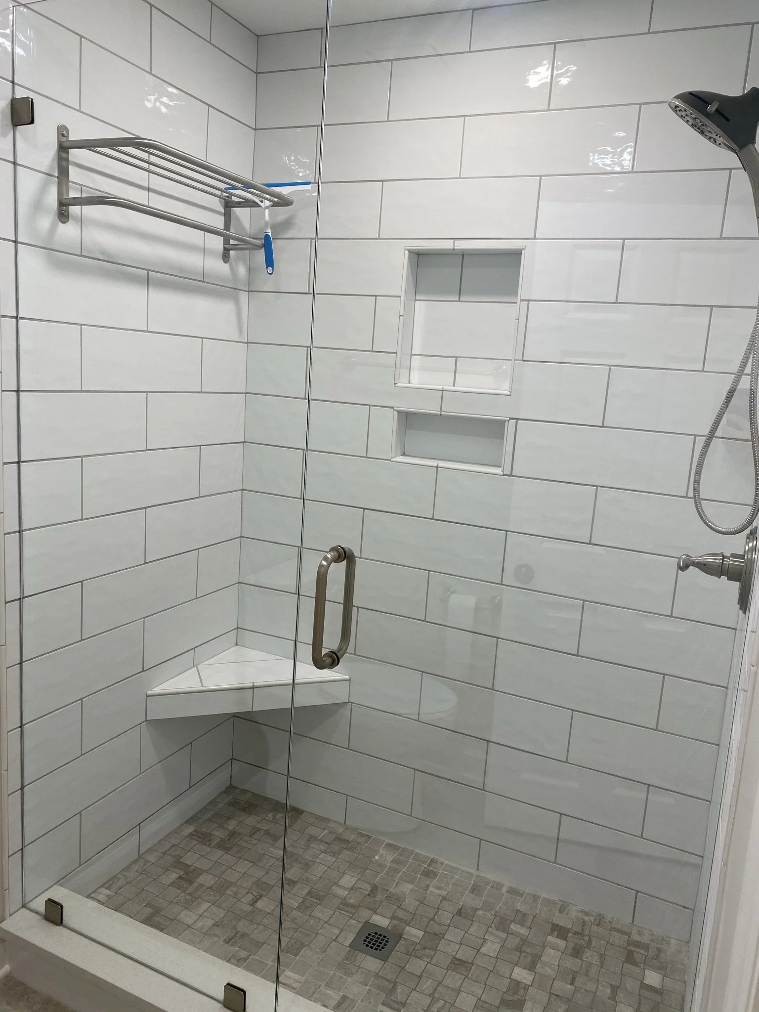 A walk-in shower with a glass door, white subway tile walls, a built-in niche with two shelves, a wall-mounted showerhead, a metal shower shelf with three bars, and a chrome grab bar inside.