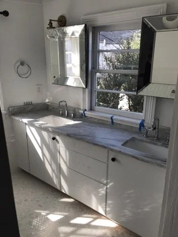 Kitchen with white cabinets and marble countertop, window with blinds, mirror, and overhead cabinet.