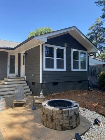 Backyard with a newly renovated gray house, outdoor seating, a stone fire pit, and a paved patio under a clear blue sky.