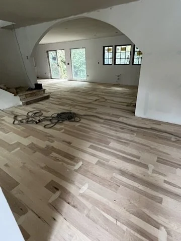 Interior of a house under renovation with hardwood flooring, an arched doorway, and large windows, some tools and construction materials on the floor.