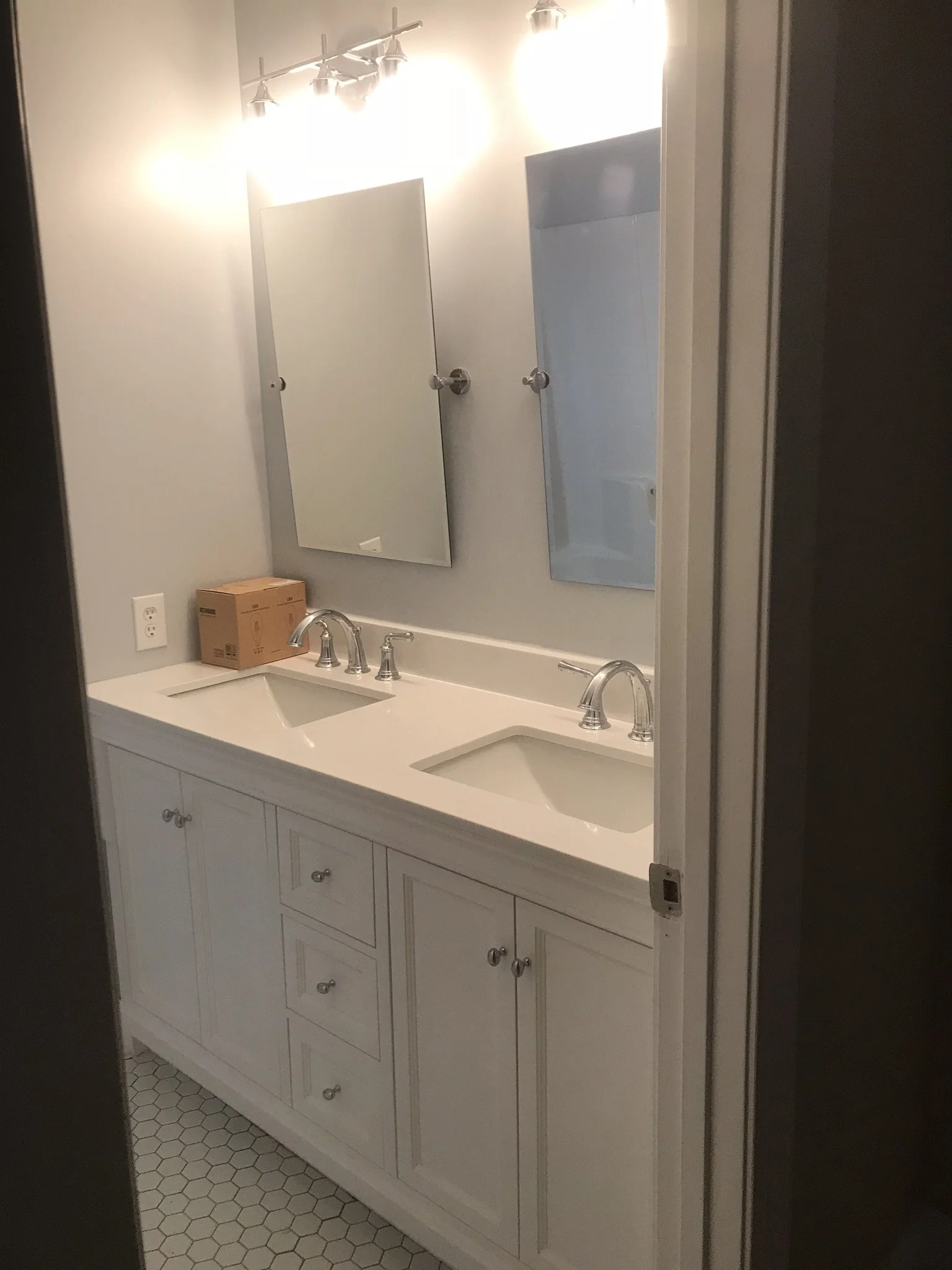Bathroom vanity with two sinks, white cabinet below, mirror, and overhead lights. A box is on the counter.