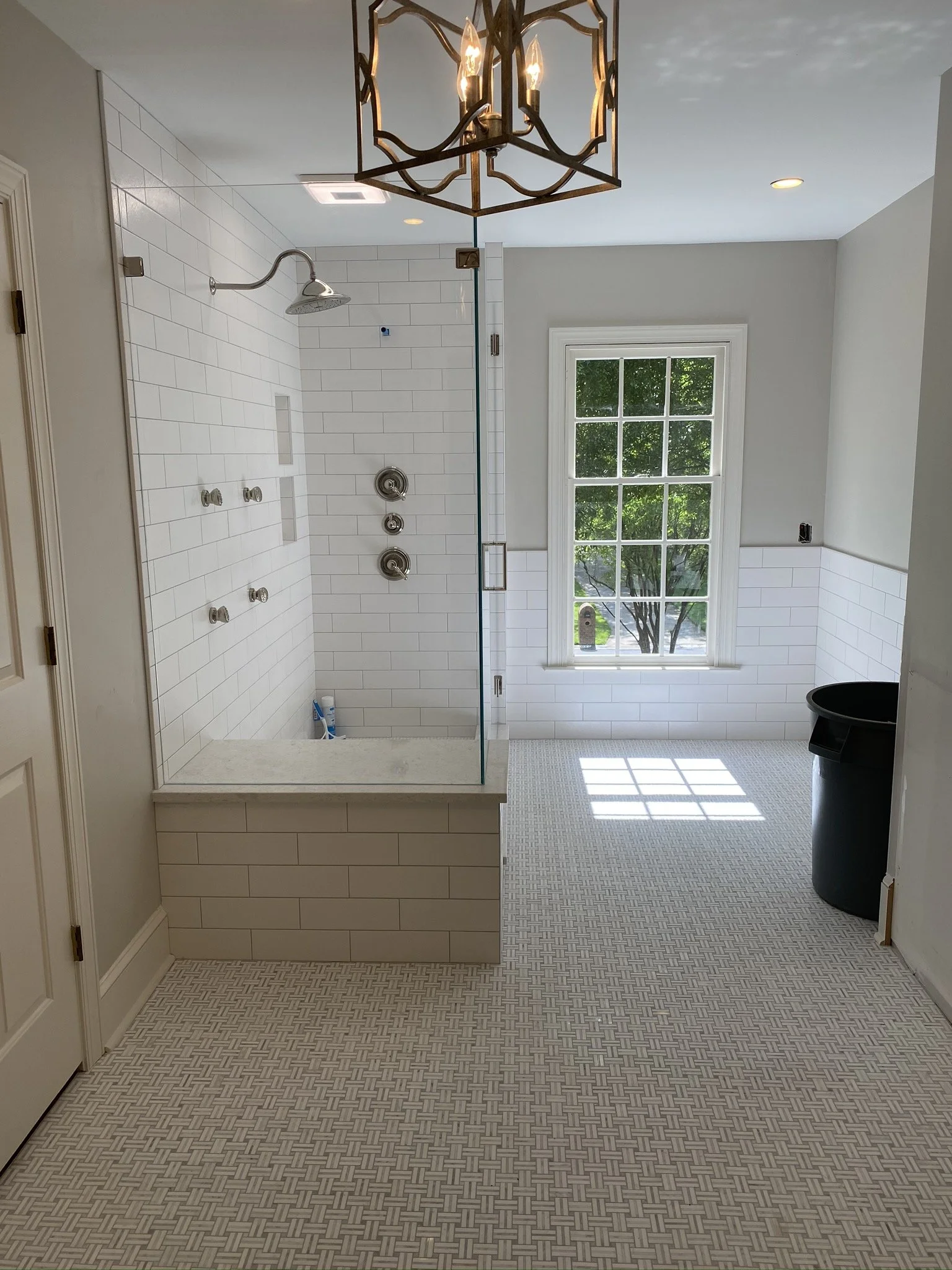 This image shows a bathroom with a walk-in shower, large window, patterned floor tiles, and a modern chandelier light fixture.