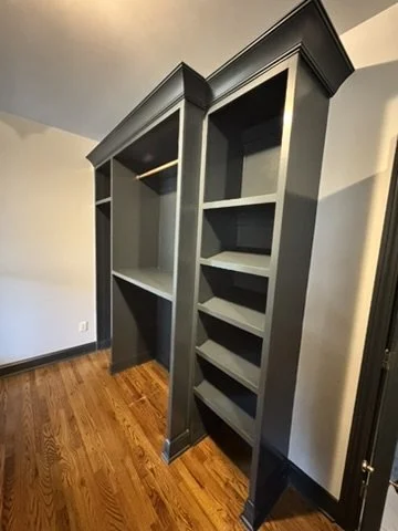 Two tall, dark gray bookshelves with multiple shelves, positioned side by side in a room with hardwood flooring and white walls.