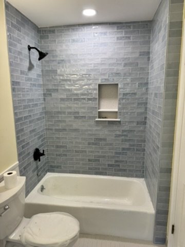 Bathroom with gray brick tile walls, a showerhead, a built-in niche, and a bathtub. Toilet with toilet paper to the left.