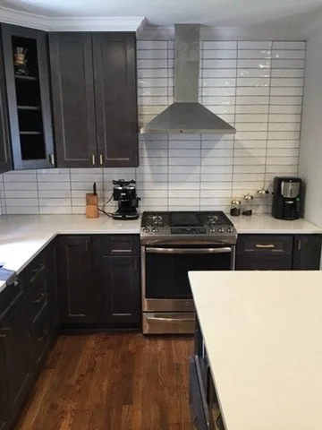 Modern kitchen with dark cabinets, white tile backsplash, stainless steel stove and vent hood, black coffee maker, and wooden floor.