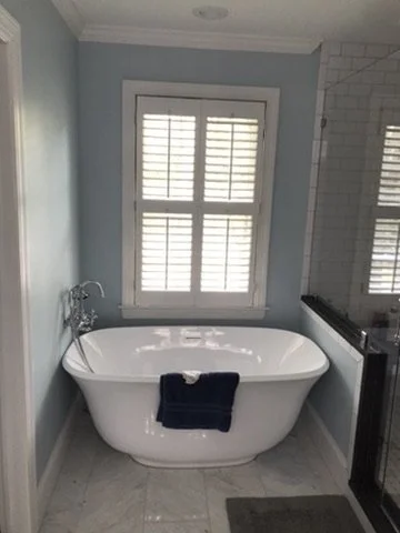 A bathroom with a white freestanding bathtub in front of a window with plantation shutters, light blue walls, and a black towel hanging on the tub.