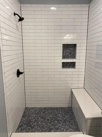 A minimalist walk-in shower with white subway tile walls, dark fixtures, a small built-in shelf, and a bench on the right side.