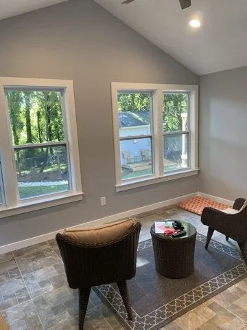 Sunlit living room with gray walls, two large windows showing greenery outside, a patterned area rug, two armchairs, a round coffee table with a book on it, and a corner bench with an orange cushion.