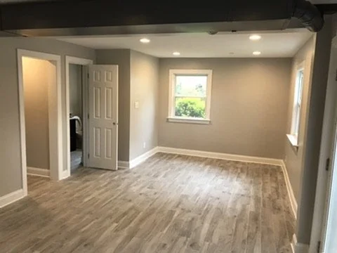 Empty living room with hardwood flooring, light gray walls, and two windows letting in natural light.