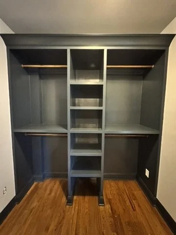 Empty built-in closet with multiple shelves and hanging rods, painted dark gray, in a room with wooden flooring.