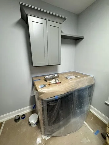 A small room with a new light gray cabinet mounted on the wall above a wrapped appliance, possibly a washer or dryer, in protective plastic. There is a small shelf on the wall to the right of the cabinet and some painting supplies on the floor.