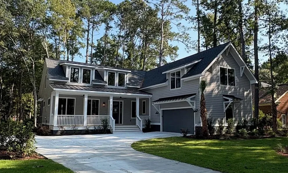 A modern two-story house with a front porch, gray siding, and a dark gray roof, surrounded by a lush green lawn and tall trees.
