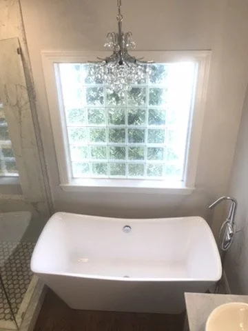 Freestanding white bathtub in a bathroom with a large window, a chandelier hanging above, and a glass shower on the left side.