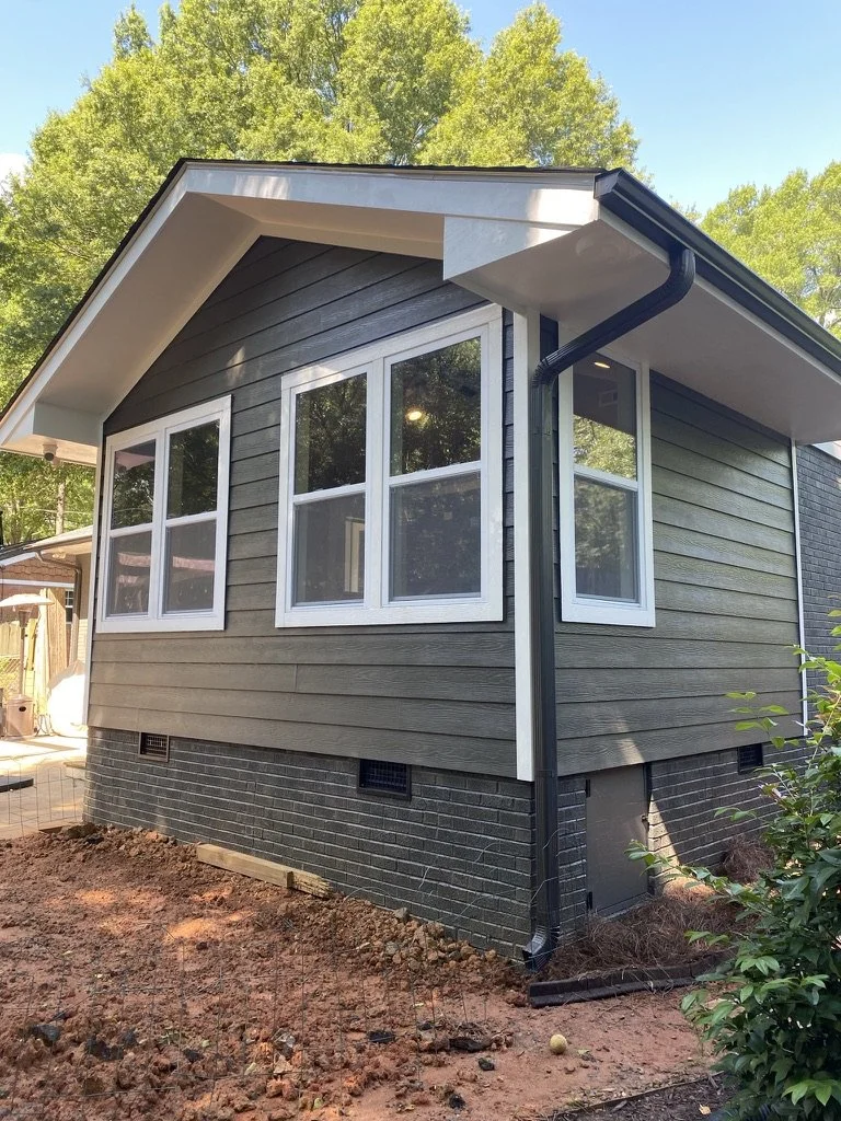 Side view of a house with gray horizontal siding, white-framed windows, black brick foundation, and black downspouts, surrounded by trees and a dirt yard.