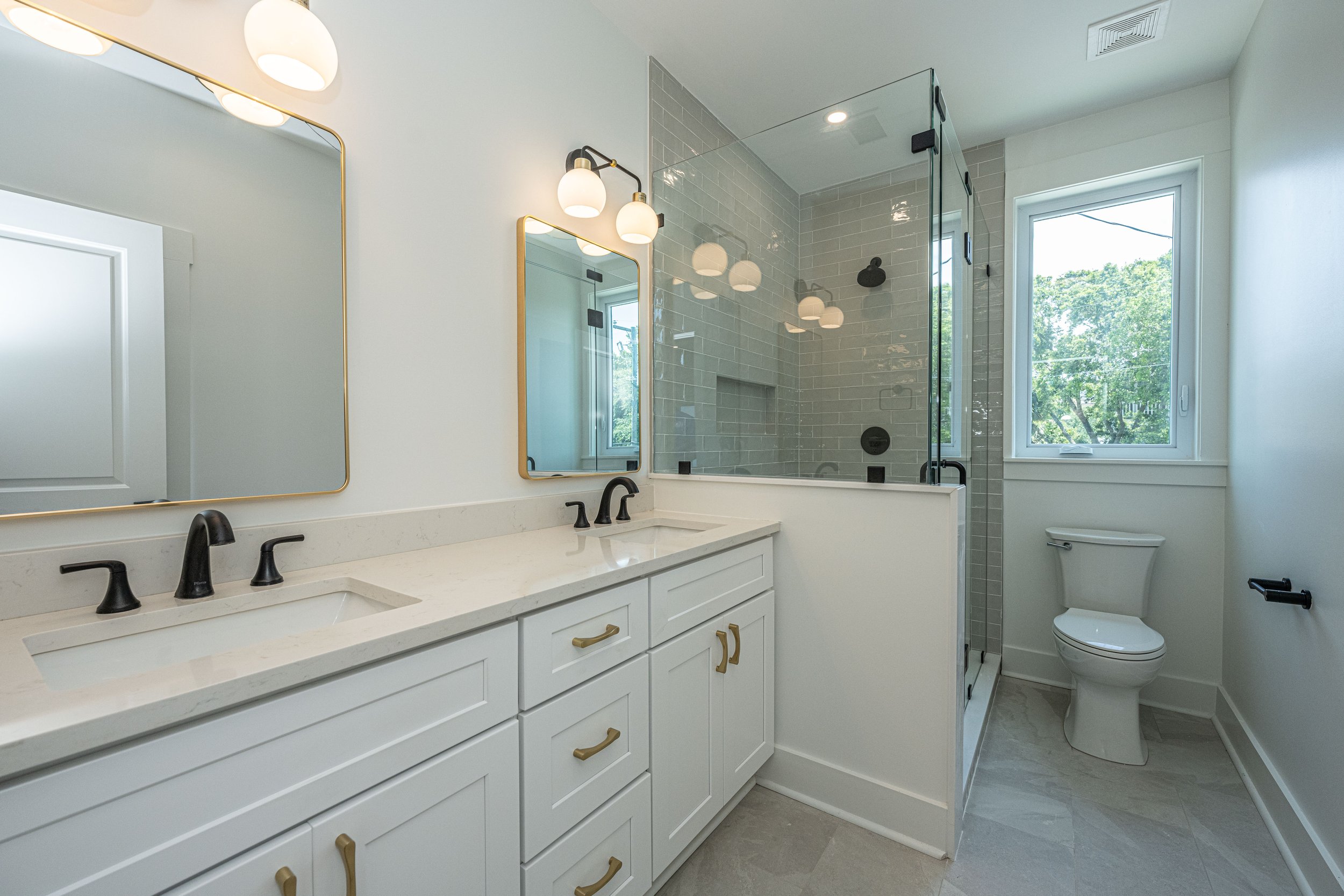 Modern bathroom with double vanity, black fixtures, large mirrors, shower with glass enclosure, and a window showing green trees outside.