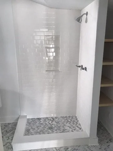 A walk-in shower with white tiled walls, a showerhead, and a built-in niche for toiletries, with a textured tile floor and a glass door.