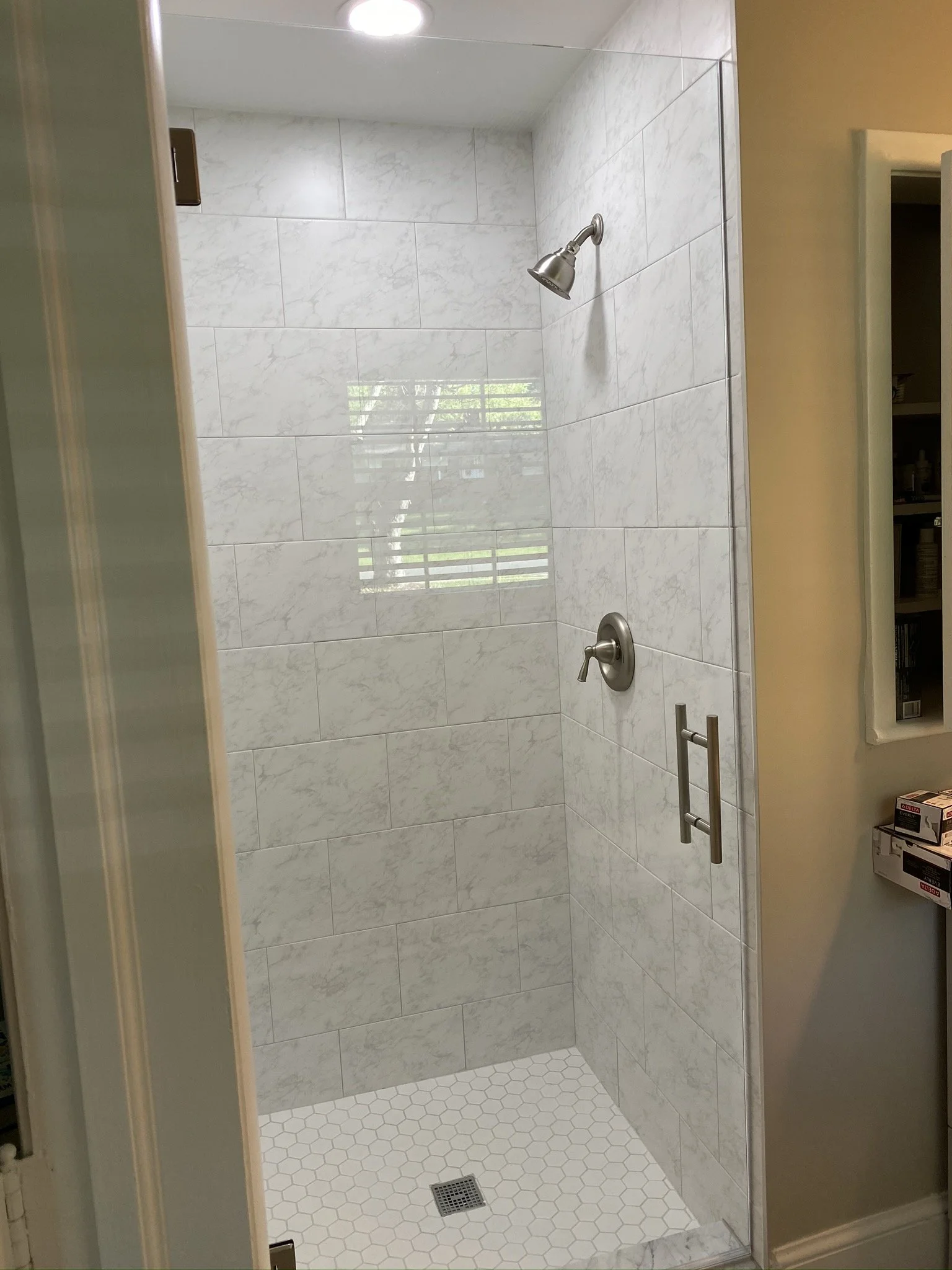 A walk-in shower with white marble tiles on the walls and a white hexagonal tile floor. There is a chrome showerhead and a valve on the wall, and a small ventilation cover on the ceiling. The shower has a partial glass door with a metal handle, and a