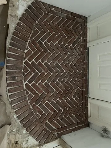 Image of a brick wall with an arched brick pattern, part of a garage or storage area, with a white door and some tools on the floor.