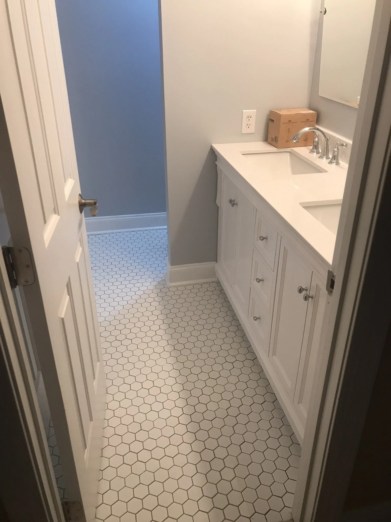 A bathroom with a white vanity with double sinks, a gray wall, a small box on the countertop, and a mirror. There's a blue wall visible through the doorway, and the floor has white hexagonal tiles.