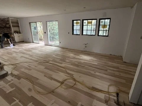 A room under construction with hardwood flooring being installed, windows, and a worker in the corner.
