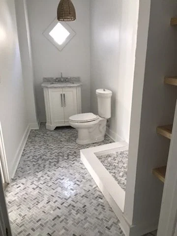 Small bathroom with a toilet, a white vanity with a sink, a mirror, and wooden shelving on the right wall.