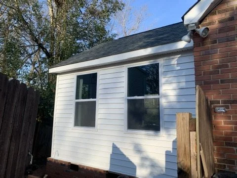 Side view of a house with white siding, two windows, and a brick wall on the right, with trees and a clear sky in the background.