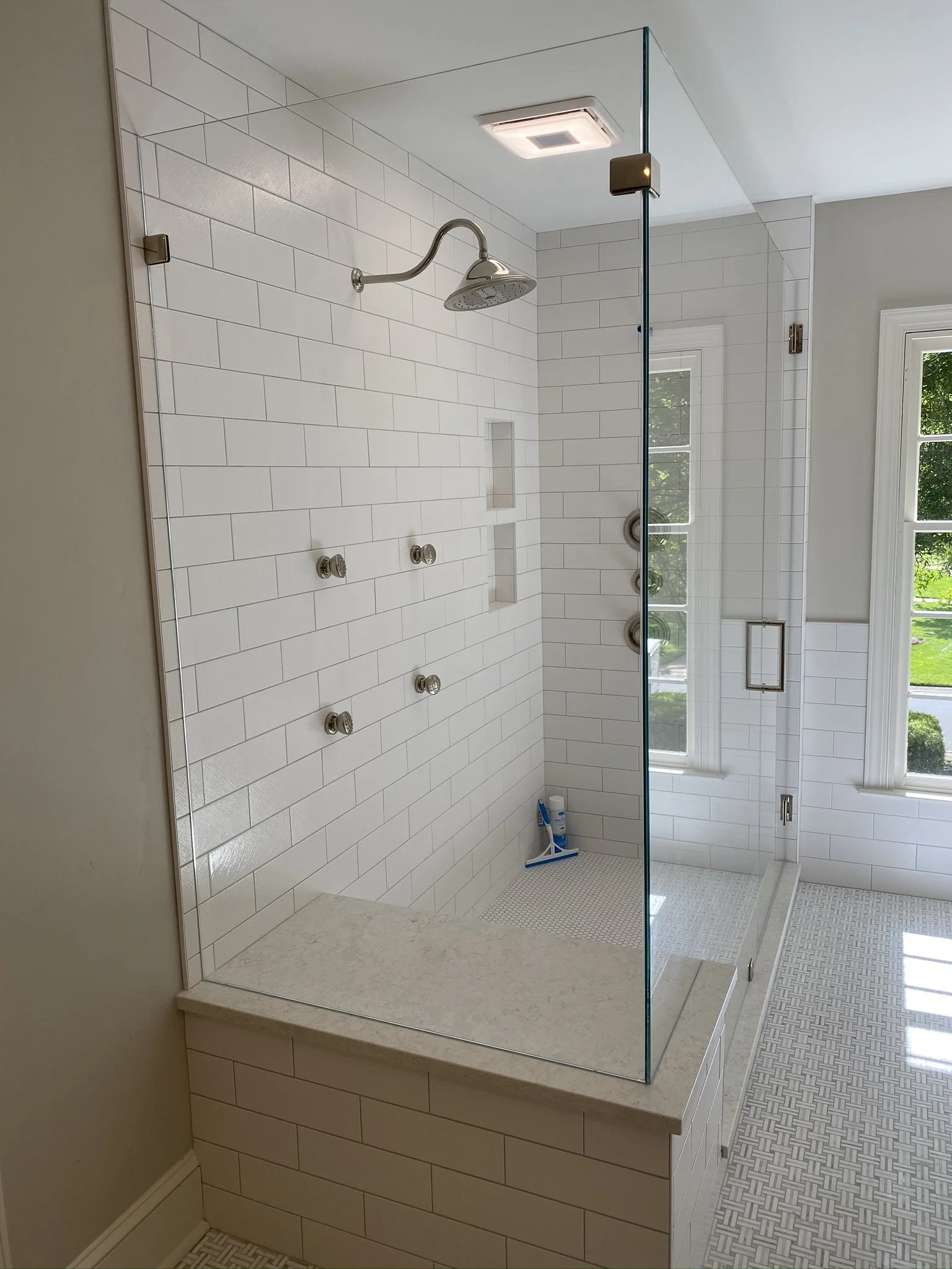 Modern bathroom shower with white subway tile walls, a rain shower head, and a glass enclosure, with sunlight coming through a window.