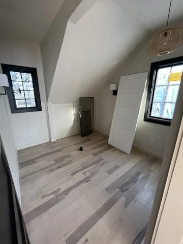 Empty attic room with light wood flooring, white walls, two black-framed windows, a white door leaning against the wall, and a ceiling light fixture.