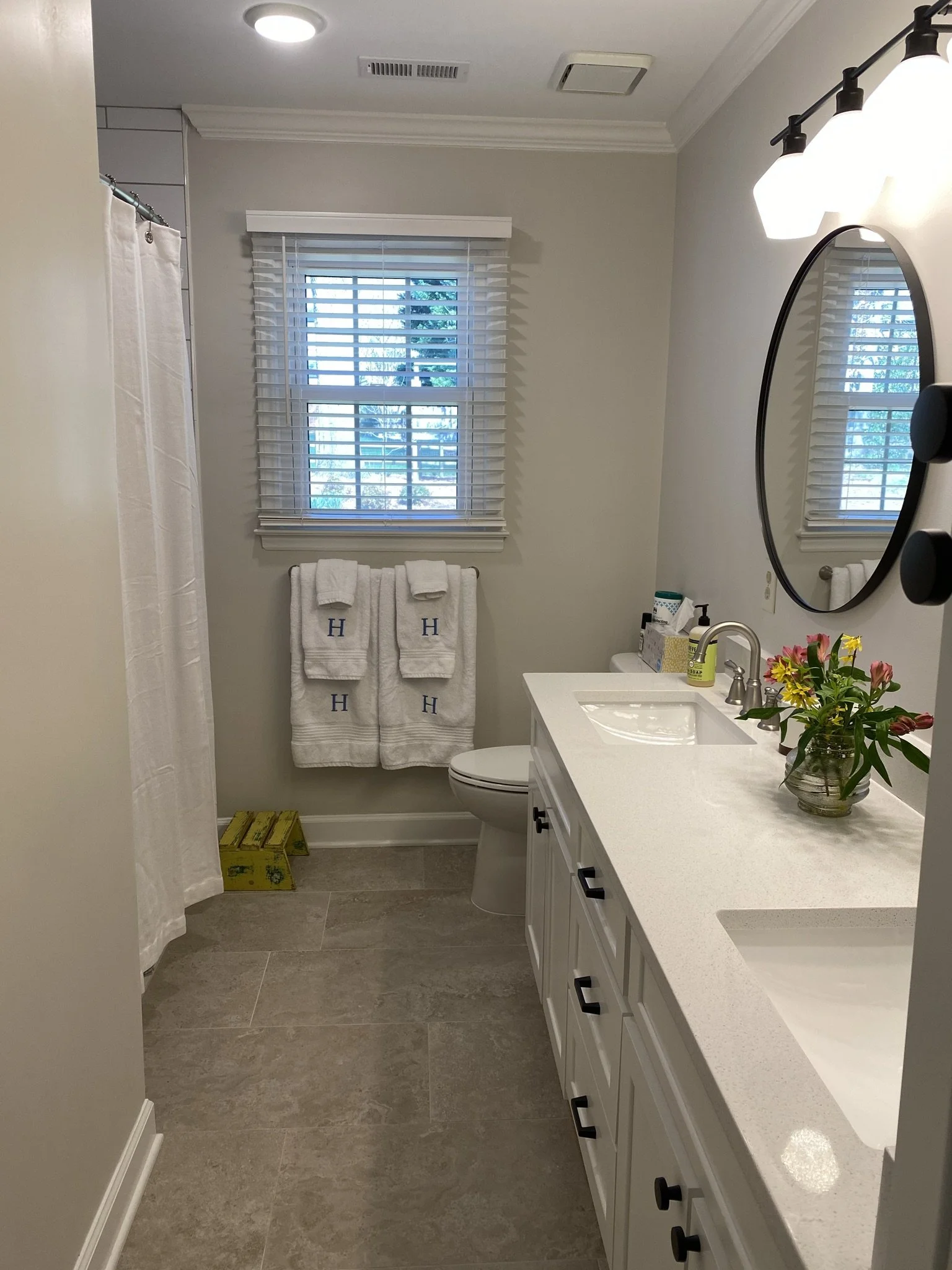 A bathroom with a vanity, a mirror, and a window with white blinds. There are towels hanging on a rack, floral decorations on the counter, and a shower curtain on the left side.